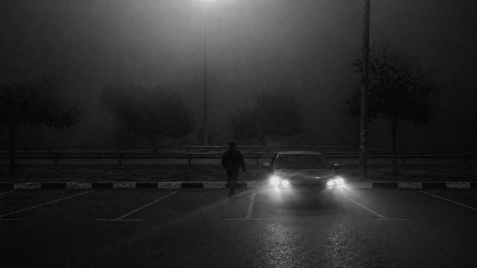 A foggy, black-and-white night scene shows a nearly empty parking lot with wet pavement reflecting light. Dim streetlights, faint trees, and a distant road fade into the mist in the background. A car is parked facing forward with its headlights on, while a lone person walks nearby in the foreground.