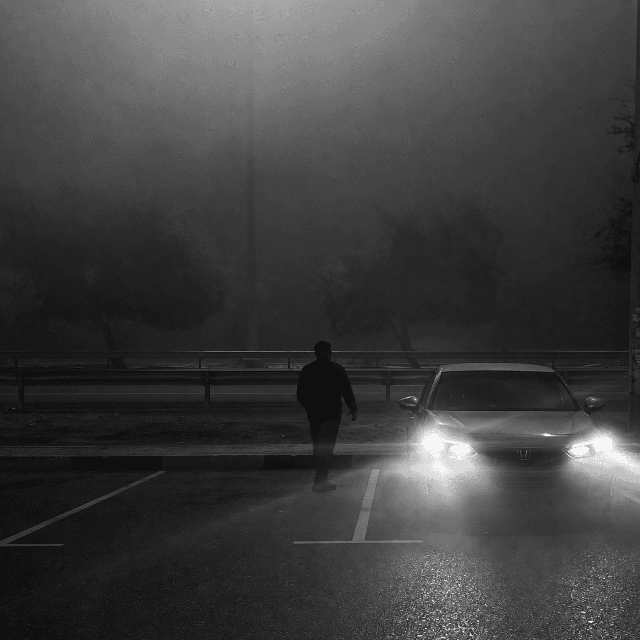 A foggy, black-and-white night scene shows a nearly empty parking lot with wet pavement reflecting light. Dim streetlights, faint trees, and a distant road fade into the mist in the background. A car is parked facing forward with its headlights on, while a lone person walks nearby in the foreground.